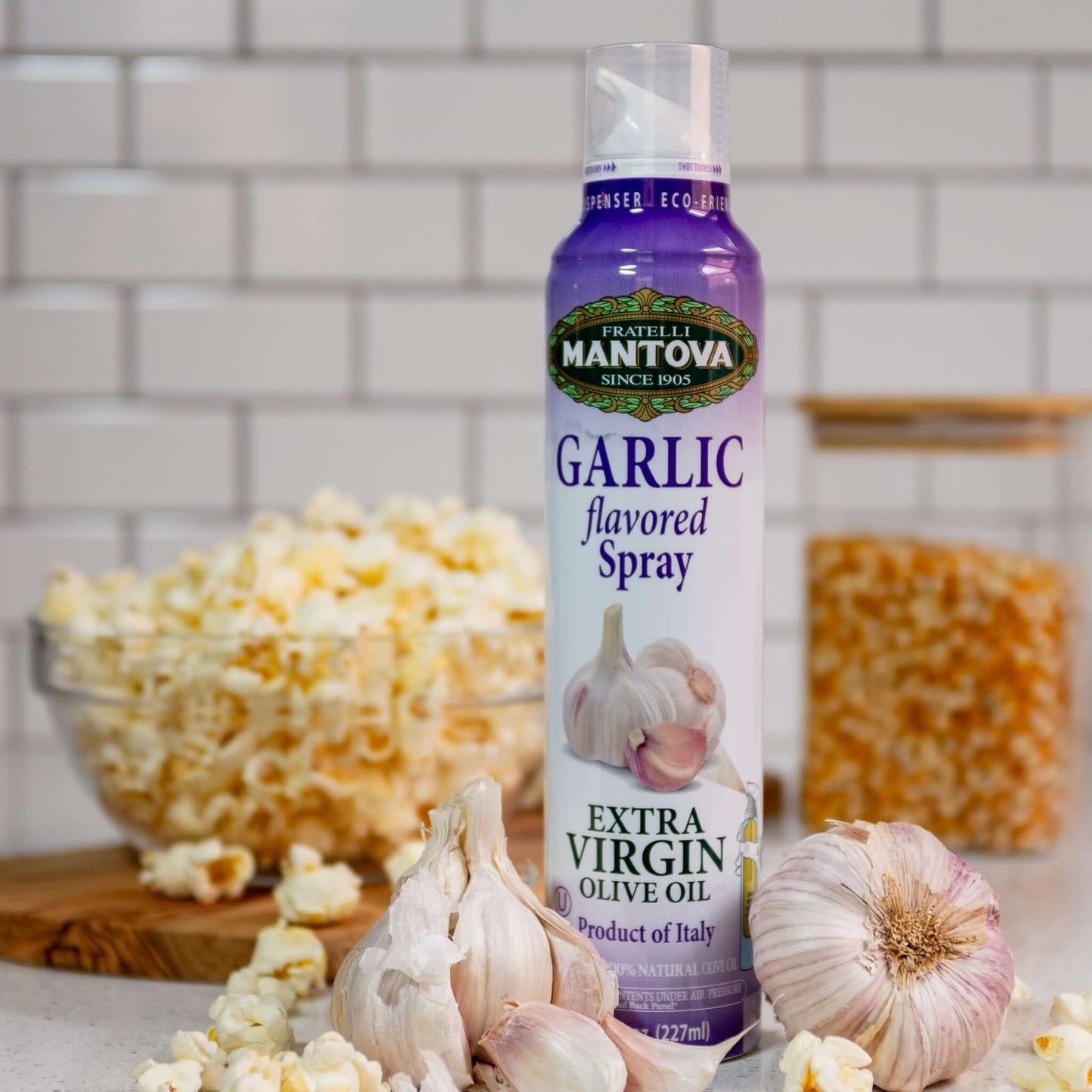 Spray bottle of garlic infused olive oil, with a head of garlic, popcorn kernels, and popped popcorn.