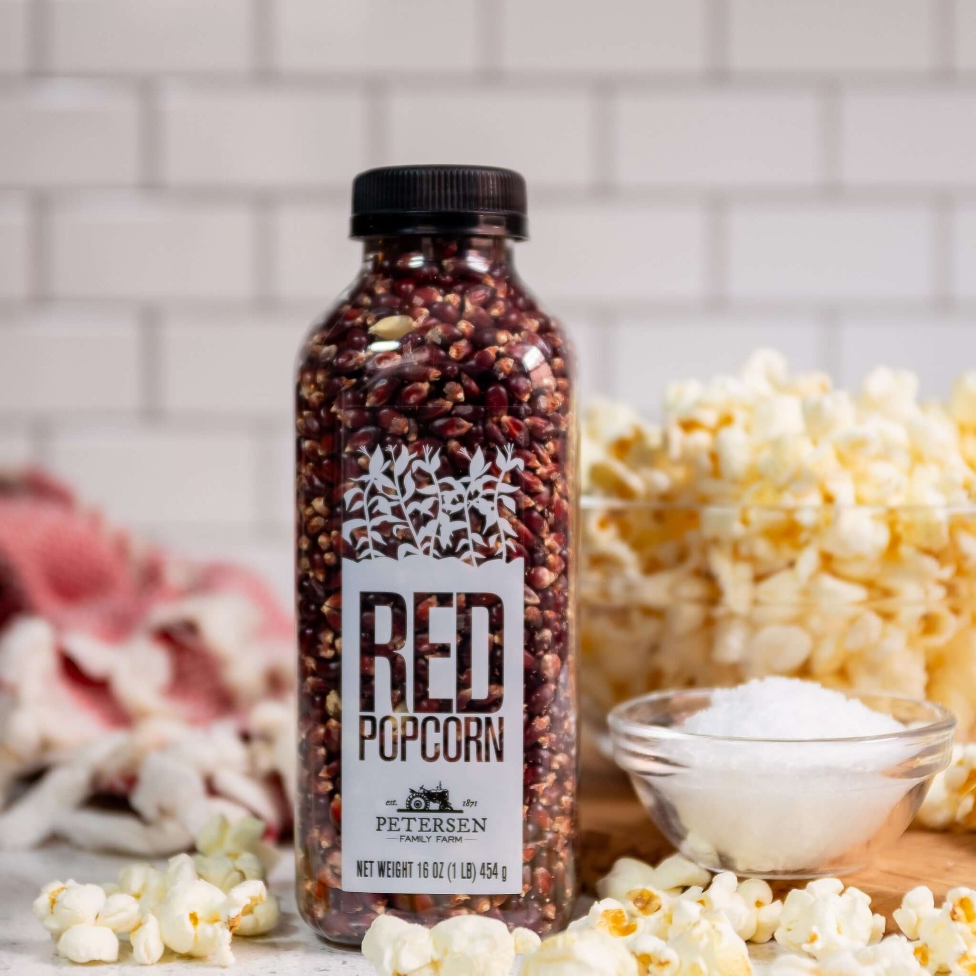 Red popcorn kernels by Petersen Family Farm. 16oz Plastic bottle on counter with popcorn