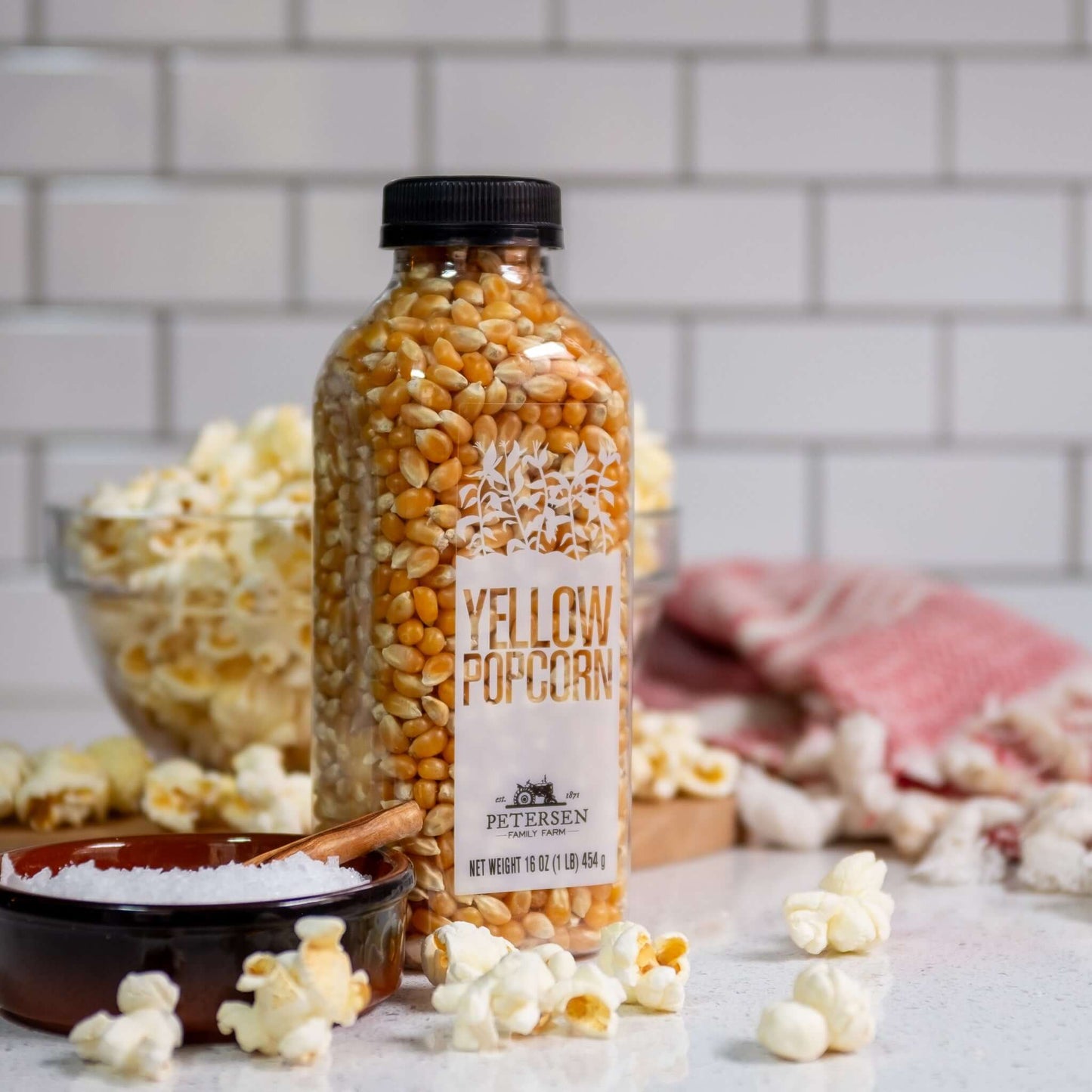 Classic yellow popcorn kernels by Petersen Family Farm. 16oz Plastic bottle, front on counter with popcorn